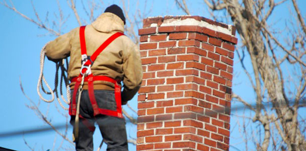 a person with their back facing the viewer standing on a rooftop next to a brick chimney-top. the person wears a coat and a harness