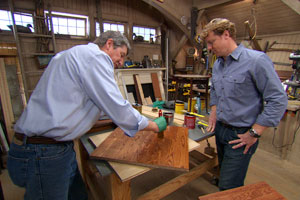 two people in a workshop with one watching the other apply finish to a piece of wood with a paintbrush